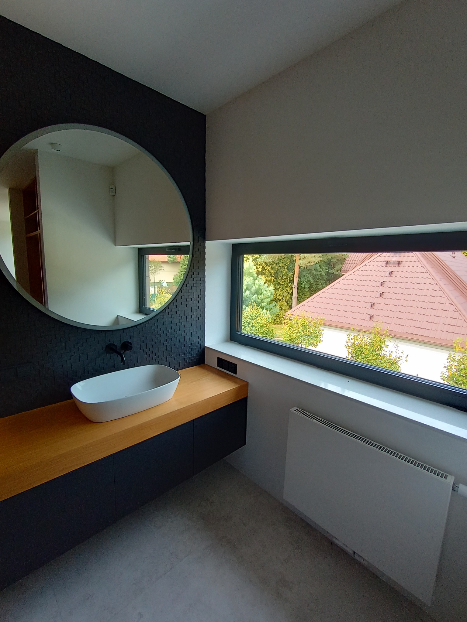 Nowoczesna łazienka z okrągłym lustrem, owalną umywalką nablatową, drewnianym blatem i czarną ścianą z dekoracyjną fakturą, widok na zewnątrz przez poziome okno.