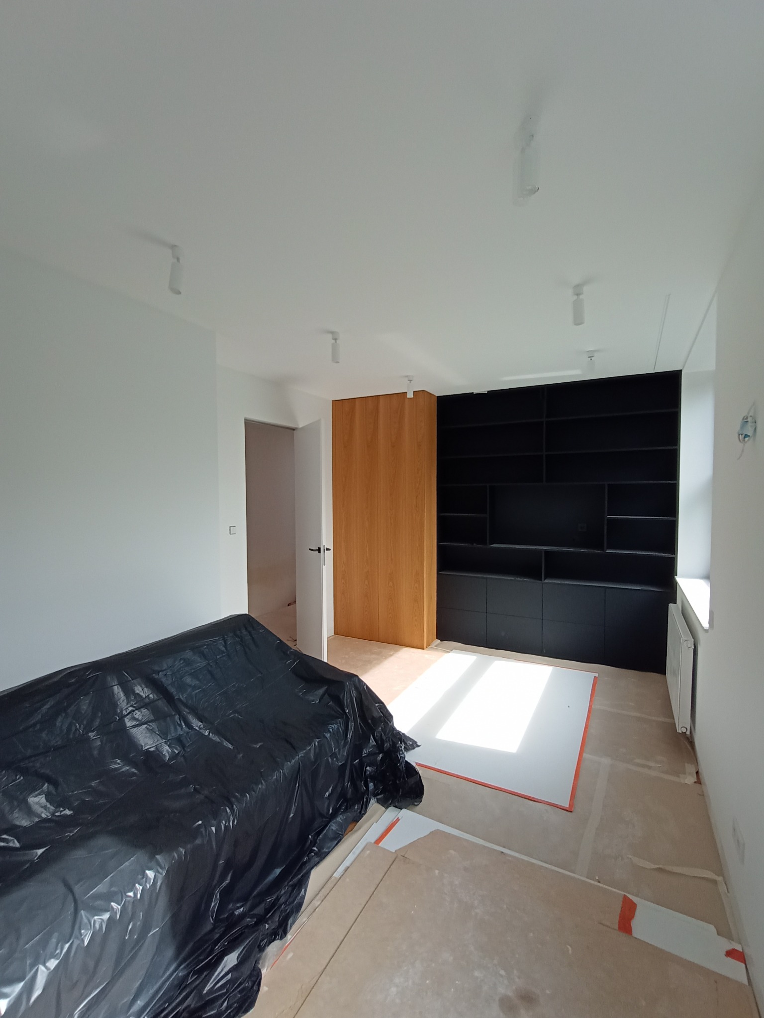 Pokój w trakcie prac w Warszawie: ściany po gładziowaniu i malowaniu na biało, podłoga zabezpieczona folią, widoczny czarny regał w zabudowie i drewniane drzwi.