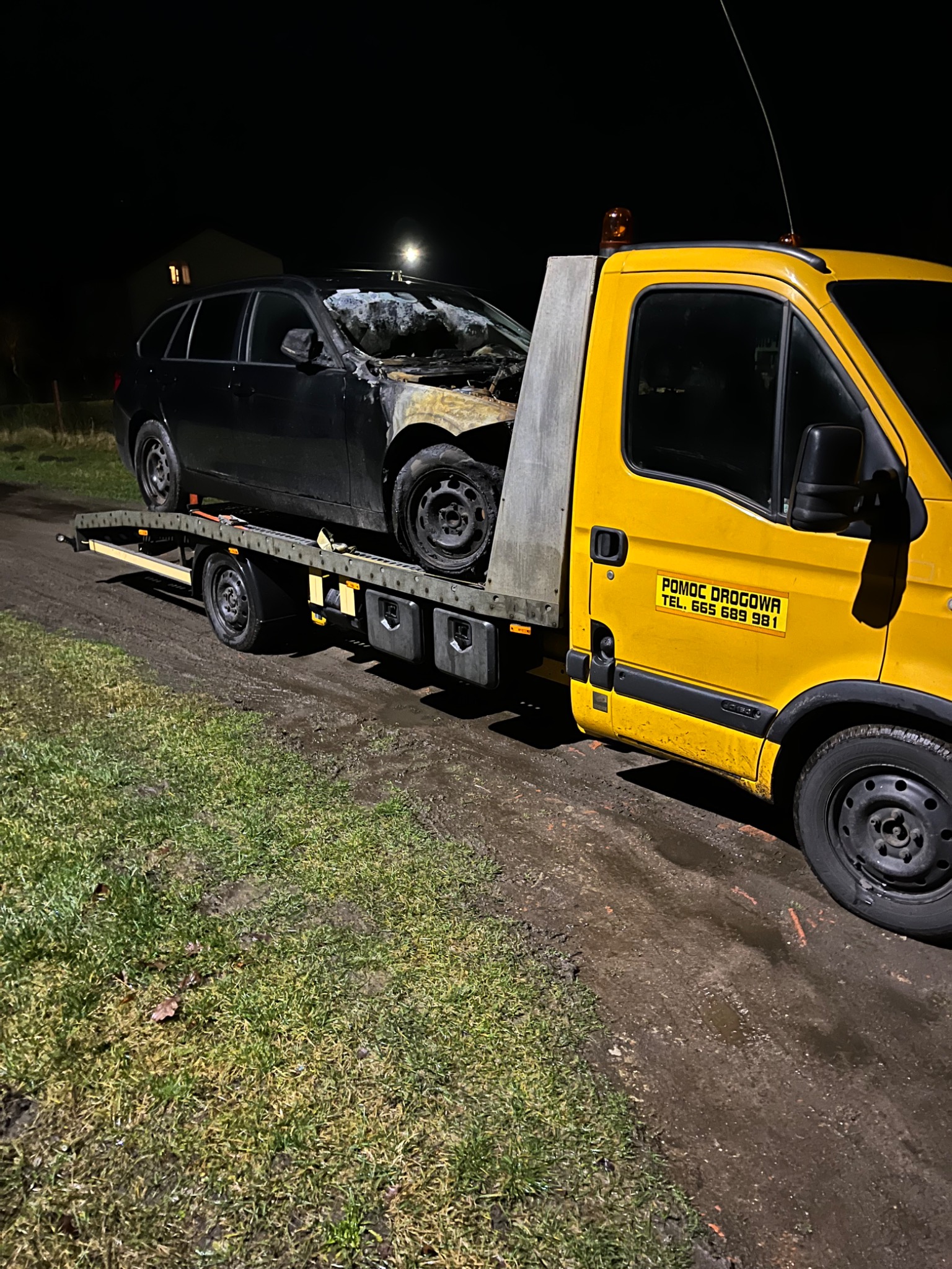 Żółta laweta z uszkodzonym, czarnym samochodem osobowym na platformie, widoczne zniszczenia po pożarze, nocne ujęcie na błotnistej drodze.
