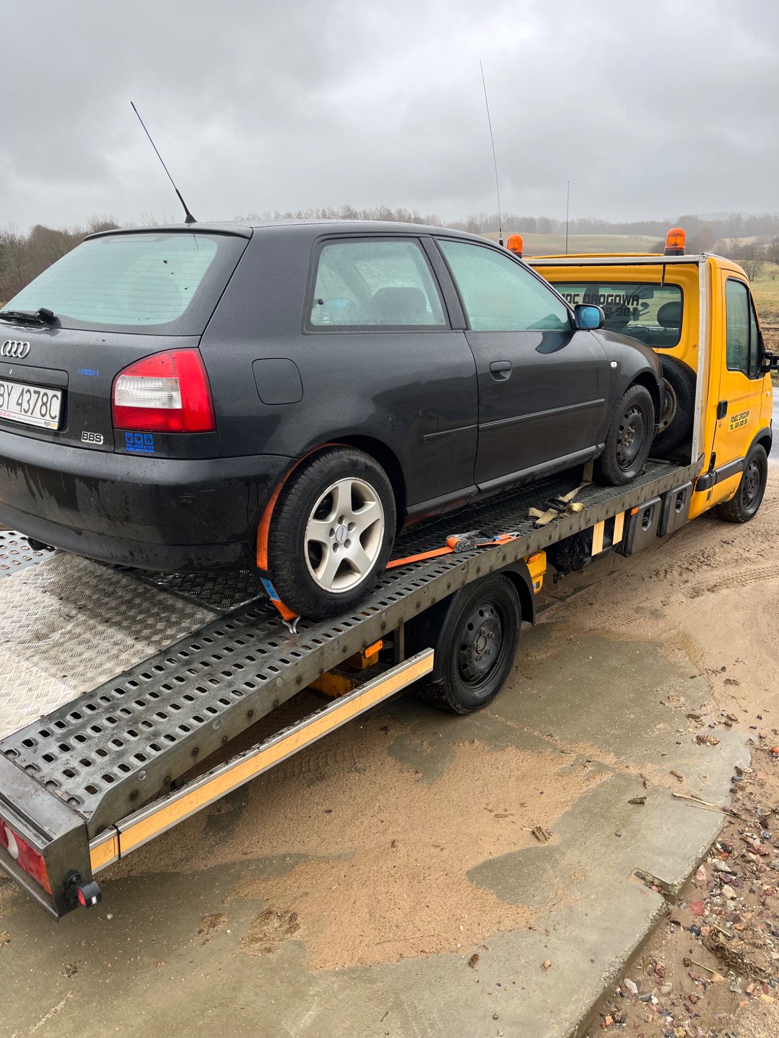 Czarny hatchback Audi A3 zabezpieczony pomarańczowymi pasami na platformie lawety w pochmurny dzień.