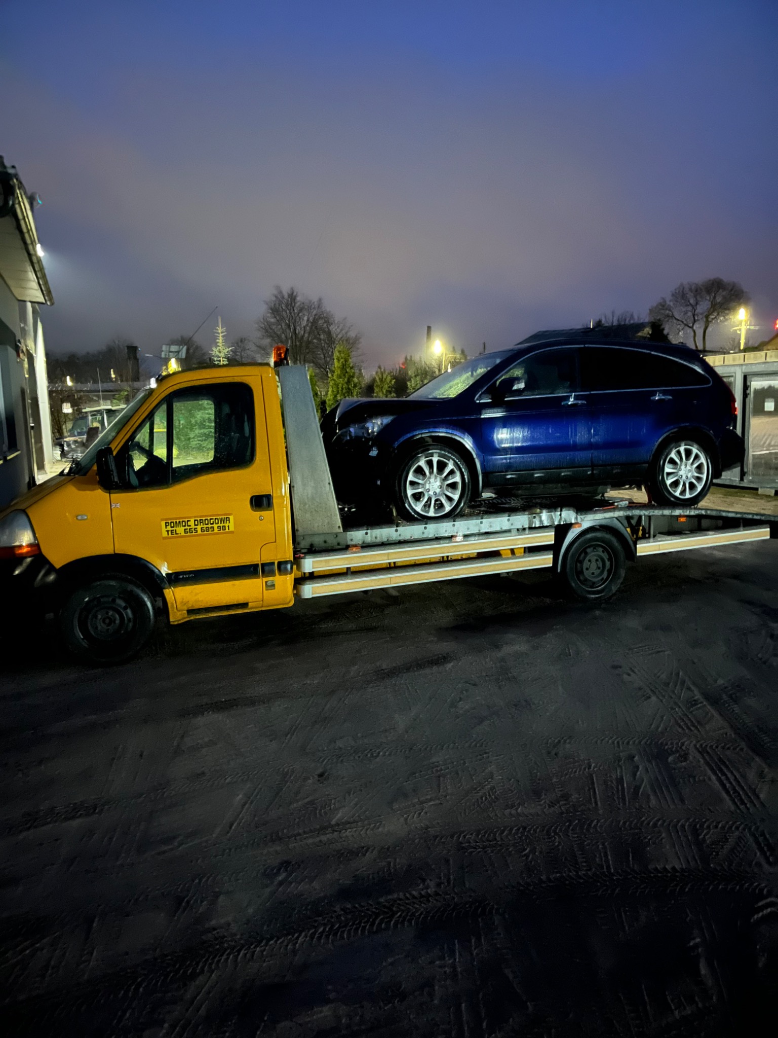 Żółta laweta z niebieskim samochodem osobowym na platformie, widoczne oznakowanie 'Pomoc Drogowa' na pojeździe, nocne ujęcie.
