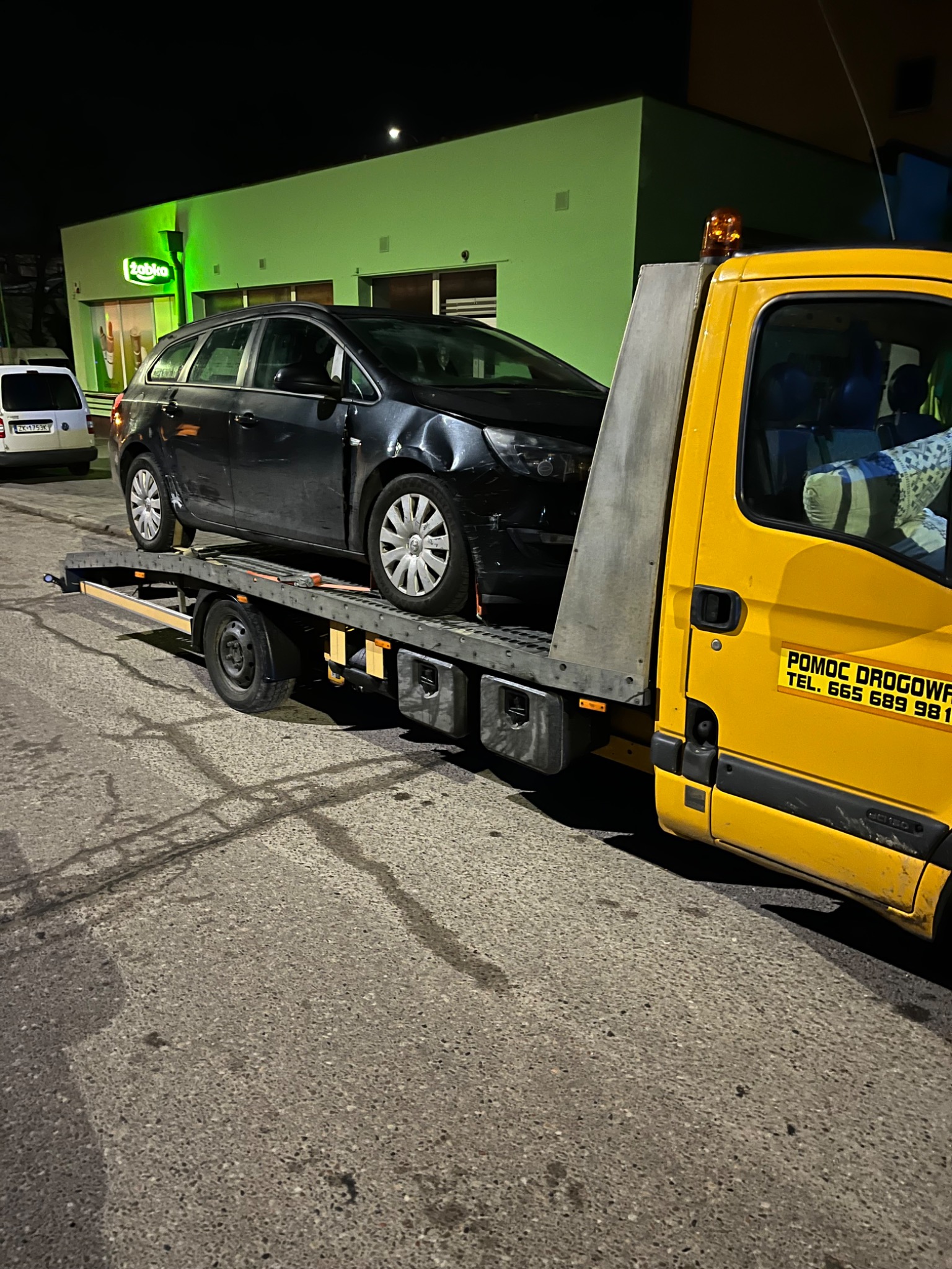 Uszkodzony, czarny samochód osobowy na żółtej lawecie pomocy drogowej, zaparkowany na ulicy w nocy przed sklepem Żabka z zielonym szyldem.