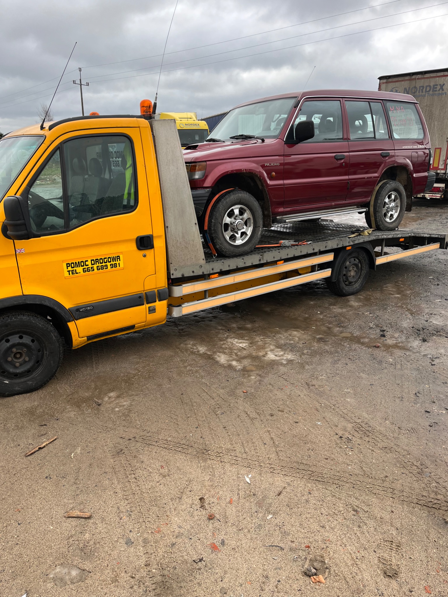 Żółta laweta z czerwonym Mitsubishi Pajero na platformie, widoczne oznaczenia 'Pomoc Drogowa' na kabinie, tło stanowi pochmurne niebo i zaparkowane pojazdy.