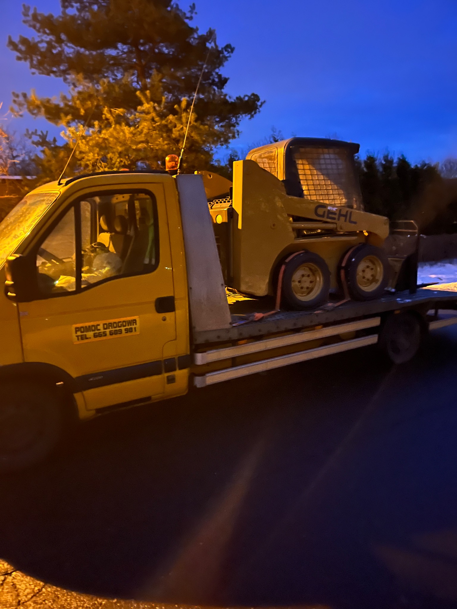 Żółta laweta z pomarańczowym kogutem przewozi żółtą miniładowarkę Gehl zabezpieczoną pasami transportowymi, na tle nocnego nieba i oświetlonego drzewa.