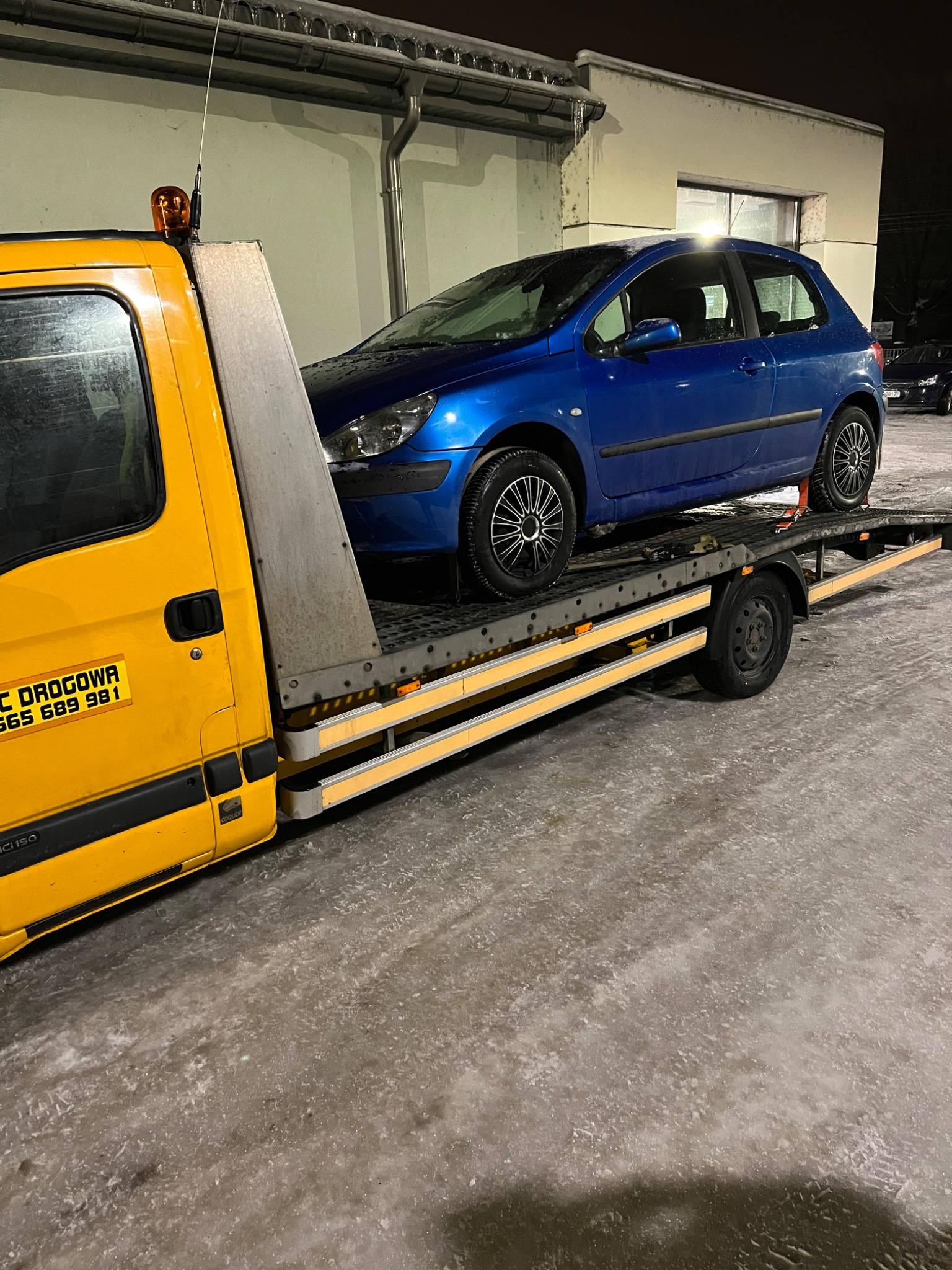 Niebieski samochód osobowy zabezpieczony pasami na platformie lawety w zimowych warunkach, widoczny żółty ciągnik z pomarańczowym światłem ostrzegawczym.