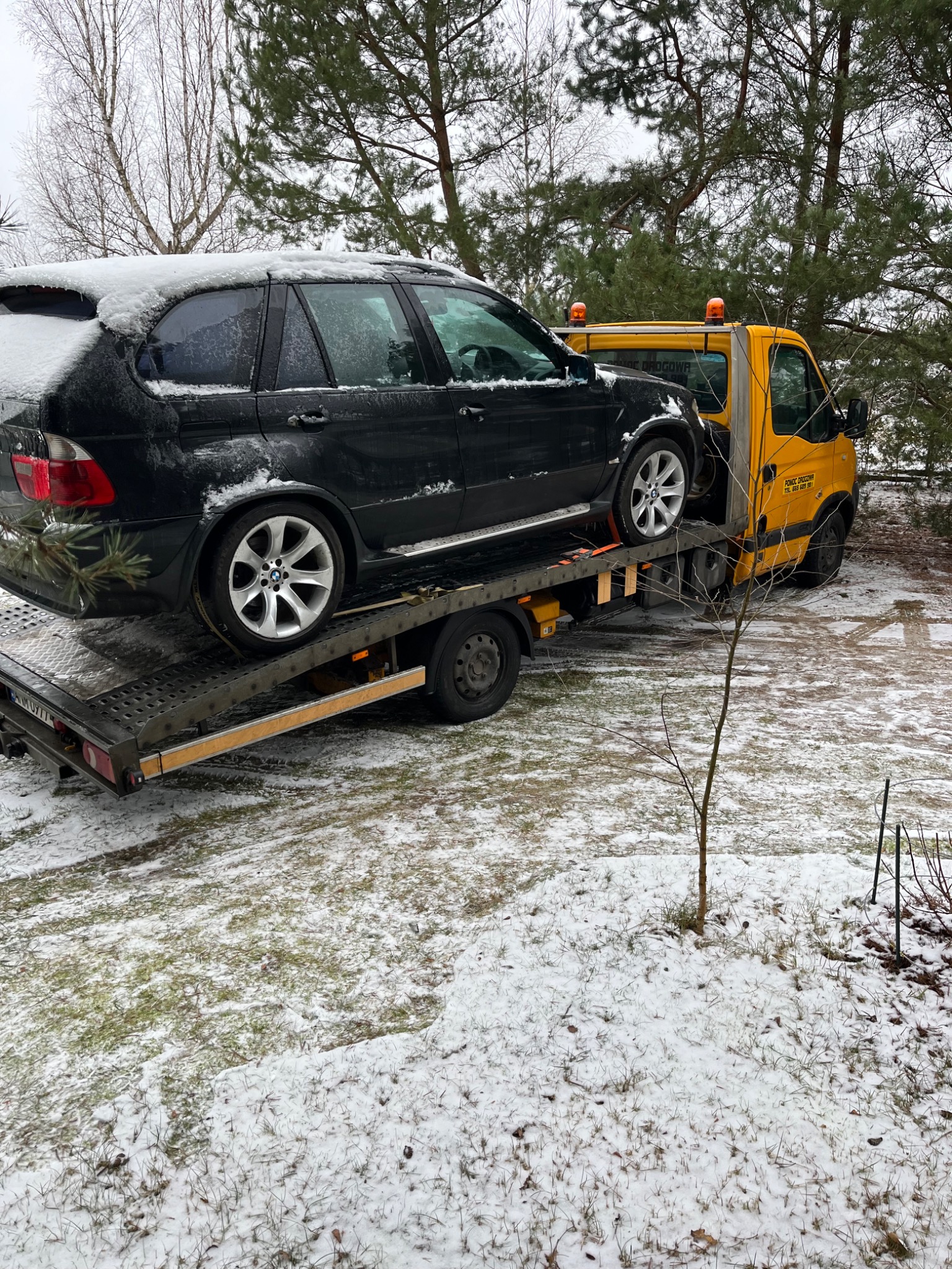 Czarny samochód typu SUV, pokryty śniegiem, przewożony na żółtej lawecie na tle zaśnieżonego terenu z drzewami.