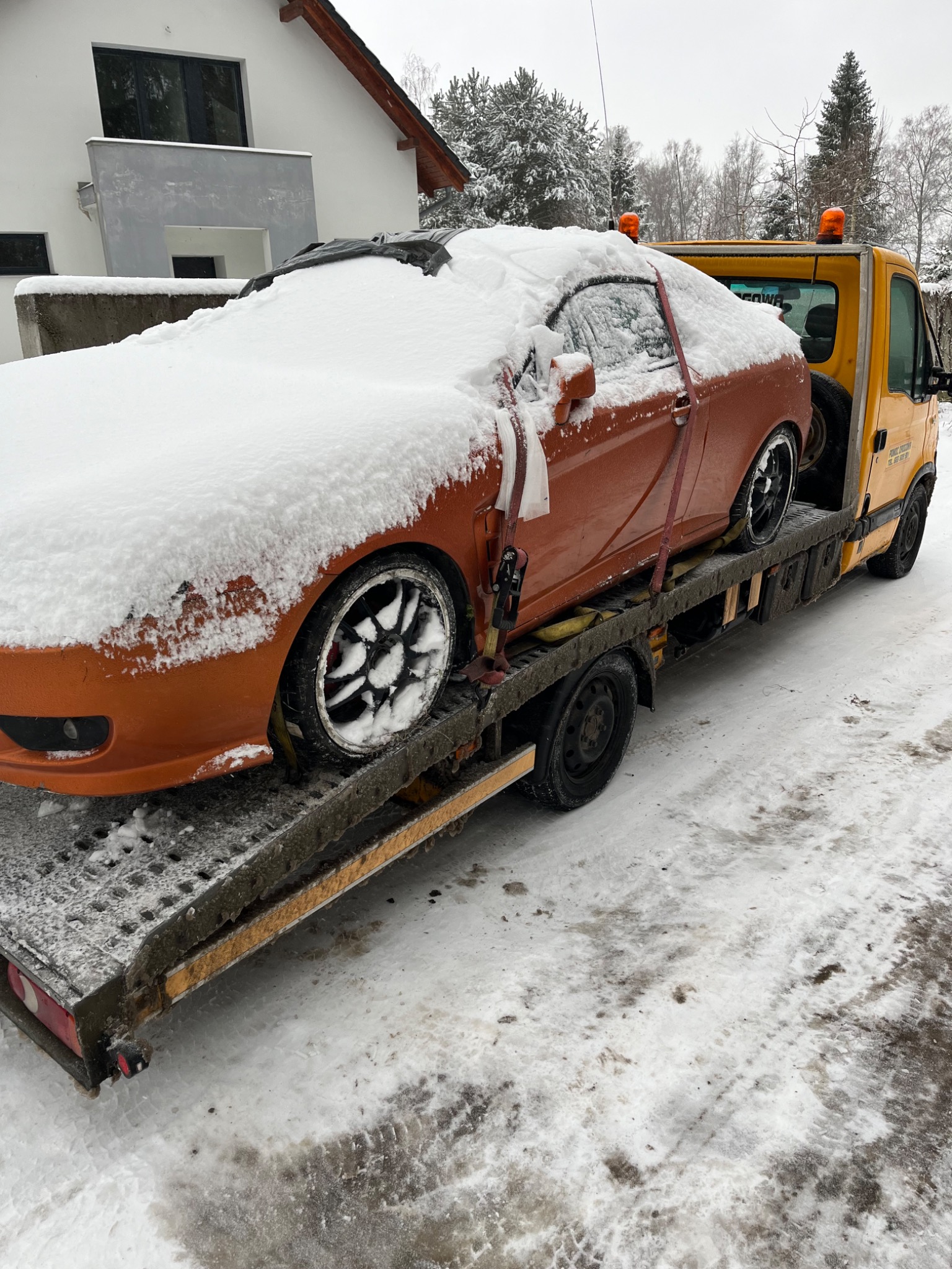 Pomarańczowy samochód sportowy częściowo przysypany śniegiem, zabezpieczony pasami na żółtej lawecie w zimowy dzień.