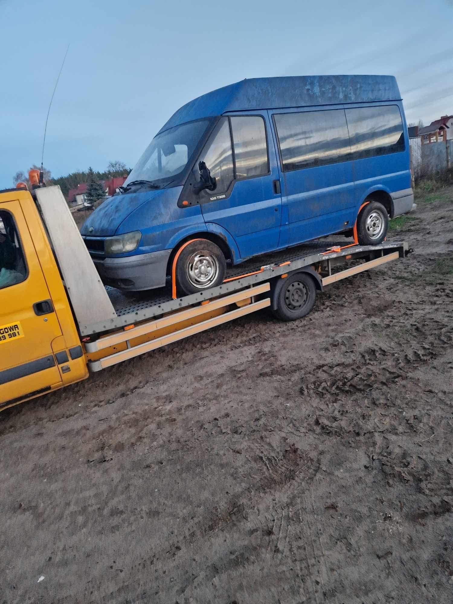 Niebieski bus Ford Transit na żółtej lawecie, zabezpieczony pomarańczowymi pasami, na błotnistym terenie.