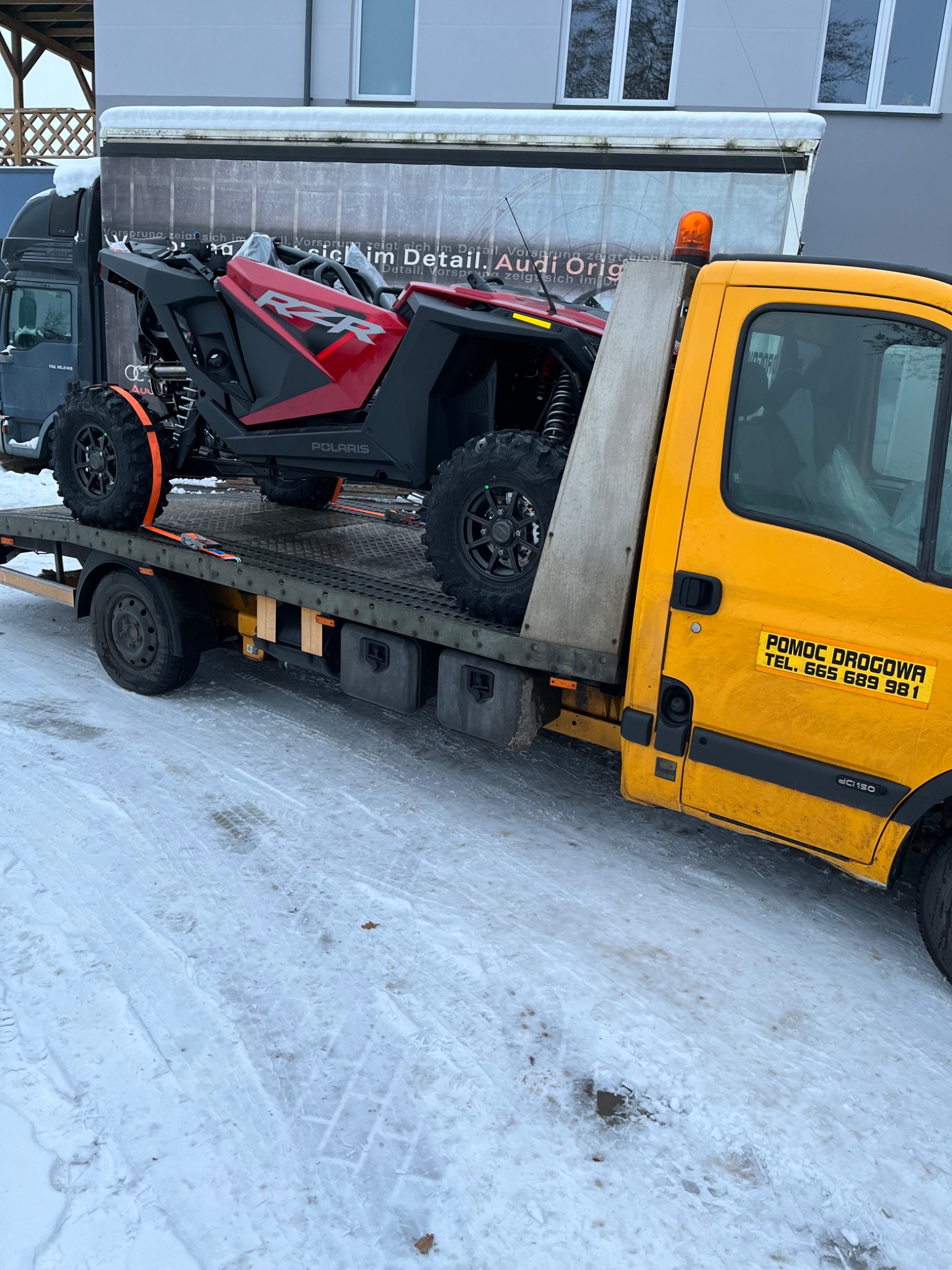 Czerwony pojazd Polaris RZR zabezpieczony pomarańczowymi pasami transportowymi na platformie lawety pomocy drogowej z żółtą kabiną i napisem 'POMOC DROGOWA' na tle zaśnieżonego terenu.