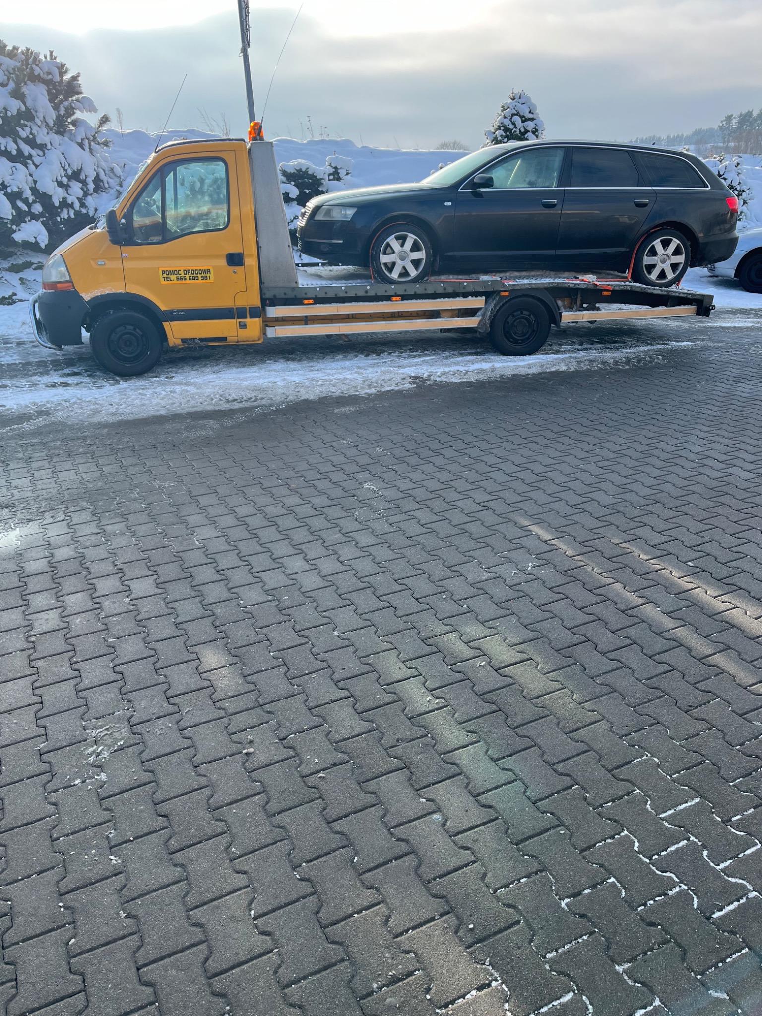 Żółta laweta z czarnym samochodem osobowym na platformie, zaparkowana na kostce brukowej w zimowej scenerii.