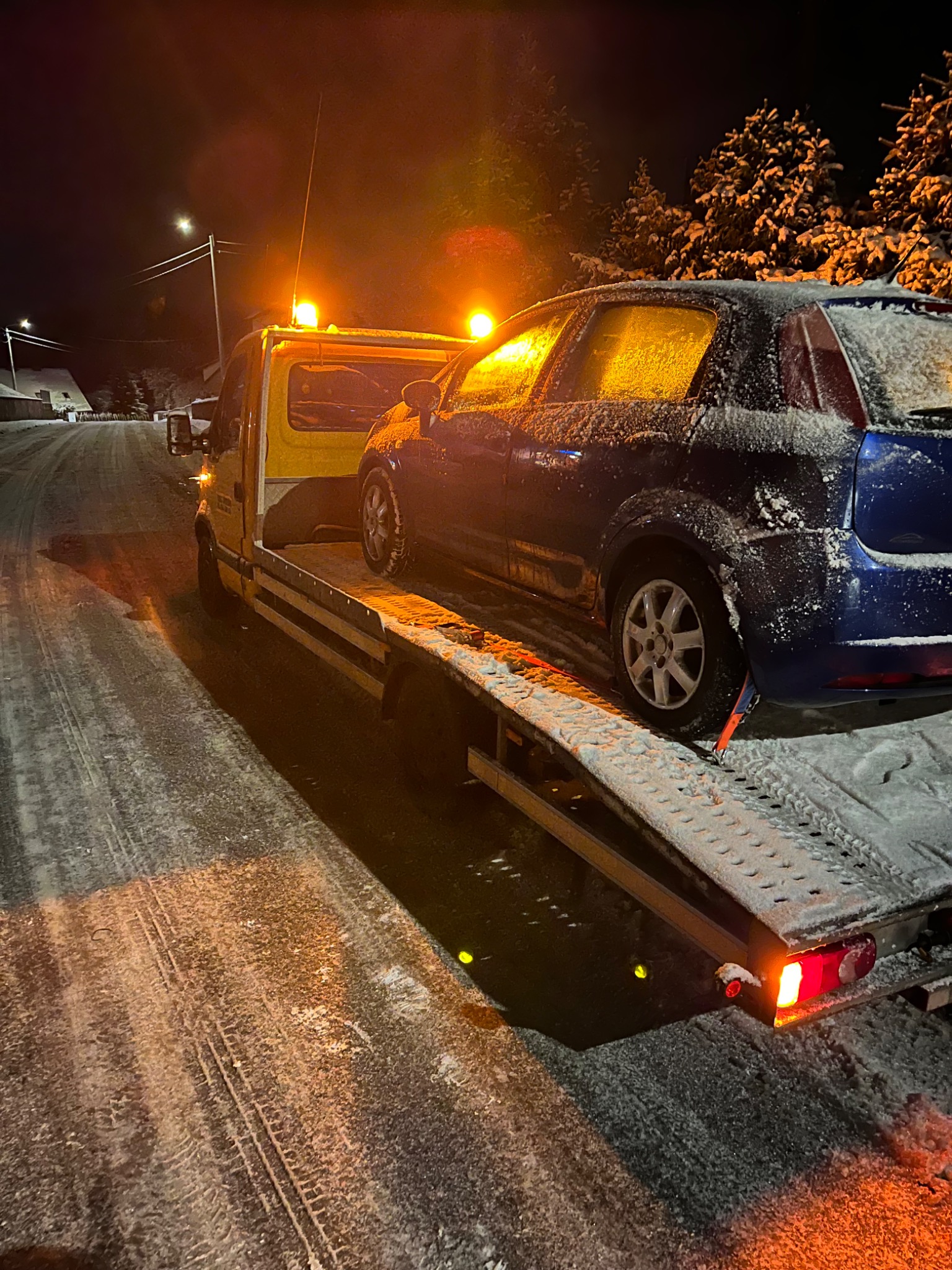 Nocna scena: niebieski samochód osobowy pokryty śniegiem, zabezpieczony pasami na platformie żółtej lawety. Śnieżna droga, oświetlenie uliczne w tle.