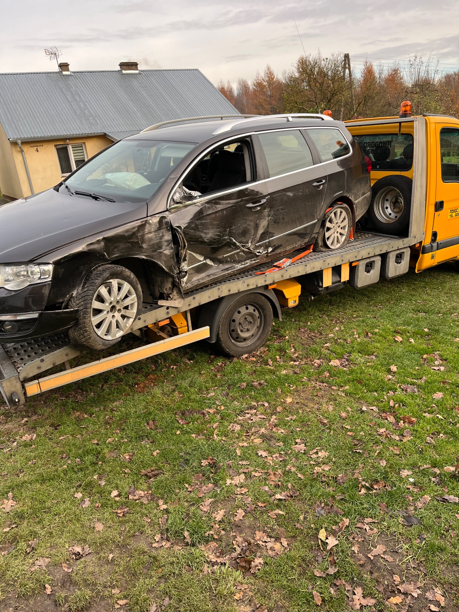 Uszkodzony czarny samochód osobowy na żółtej lawecie w jesiennej scenerii, widoczne poduszki powietrzne i uszkodzenia karoserii.