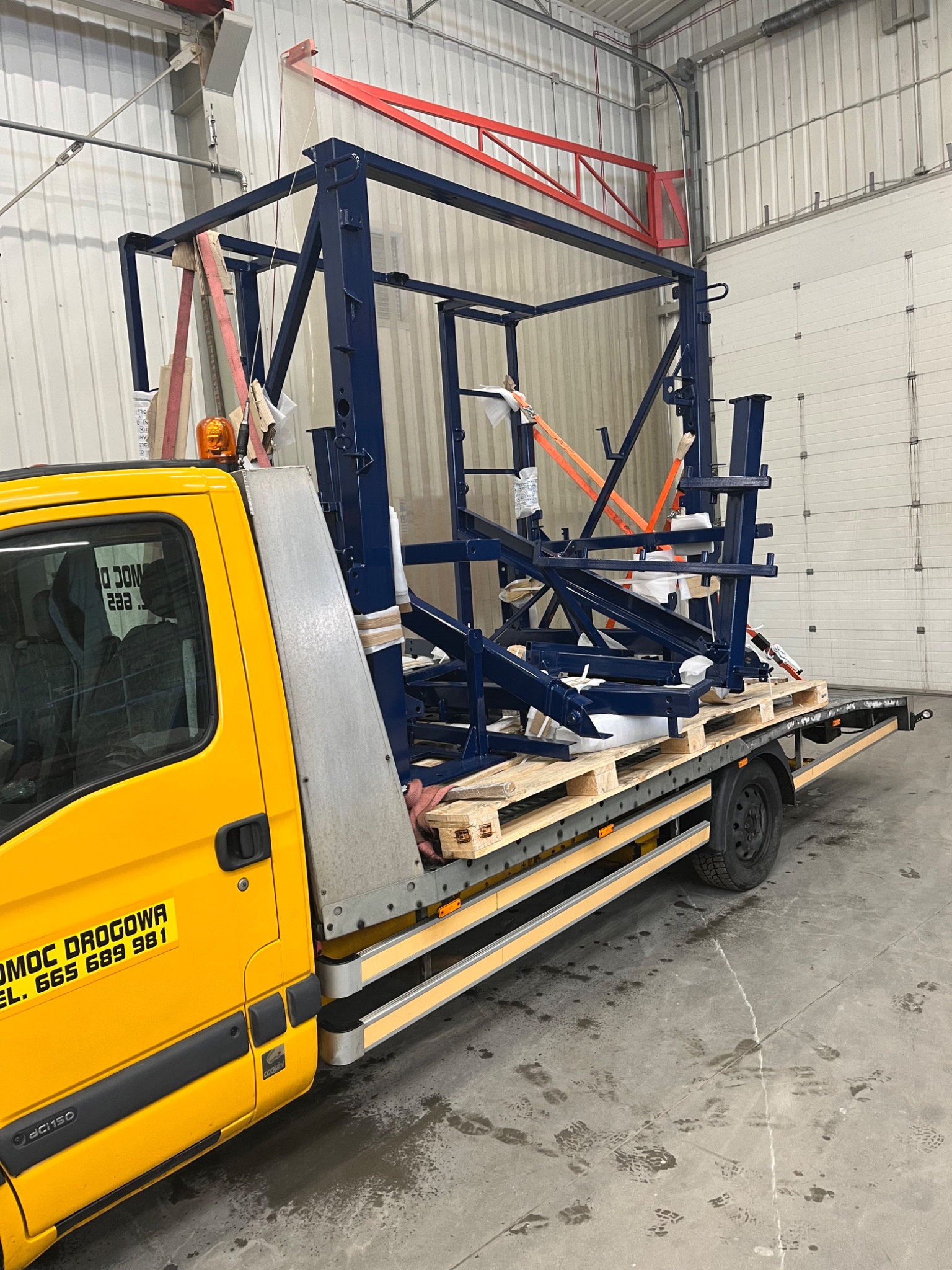Żółta laweta z konstrukcją stalową w kolorze granatowym, zabezpieczoną pasami transportowymi na paletach, transportowana w hali.