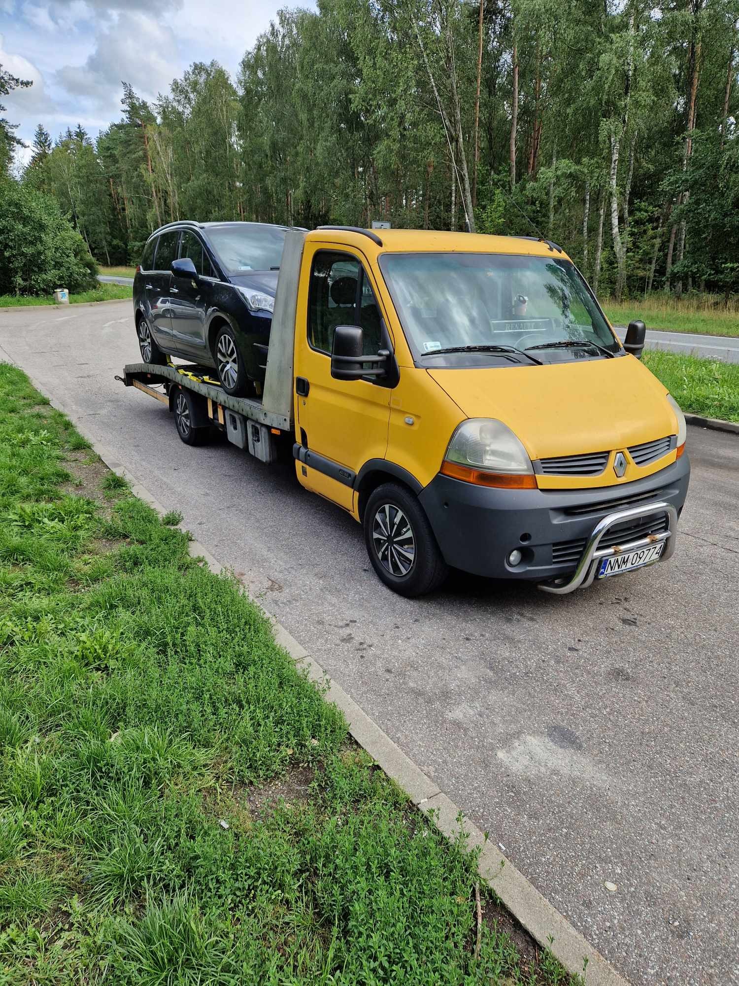 Żółta laweta Renault z załadowanym granatowym minivanem na poboczu drogi, las w tle.