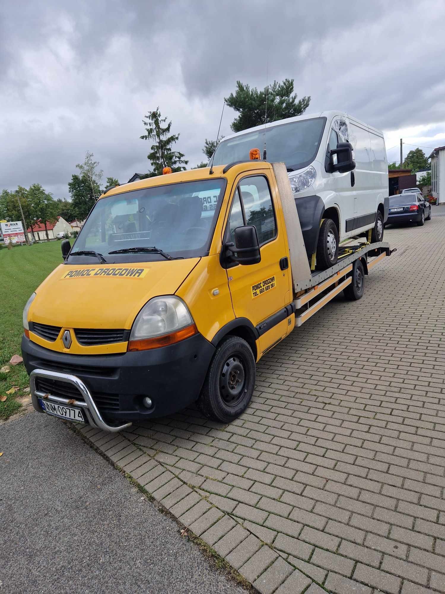 Żółty samochód pomocy drogowej z białym busem na platformie, zaparkowany na chodniku z kostki brukowej.