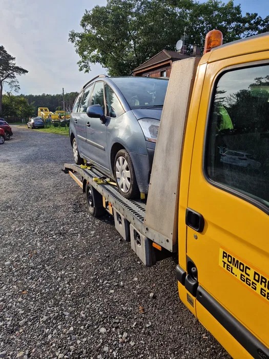 Niebieski samochód osobowy, zabezpieczony pasami, na platformie żółtej lawety z napisem 'Pomoc Drogowa', na żwirowym parkingu.