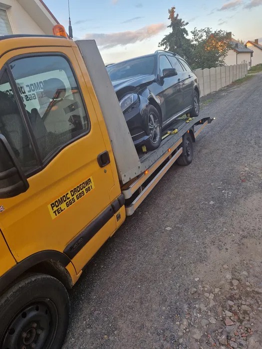 Żółta laweta z uszkodzonym, czarnym samochodem osobowym na platformie, widoczne zabezpieczenia pasami.