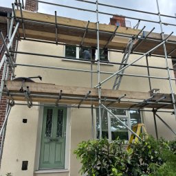 KD BUILDING - Dom jednorodzinny w rusztowaniach po nałożeniu beżowego tynku. Widoczna zielona stolarka okienna i drzwiowa. Prace elewacyjne w toku, osłonięte roślinnością.