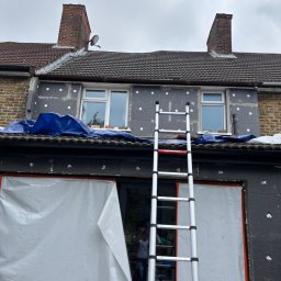 KD BUILDING - Ocieplenie elewacji domu jednorodzinnego z użyciem styropianu grafitowego. Widoczna drabina oparta o ścianę, zabezpieczona folią okienna stolarka. Etap przed nałożeniem tynku.