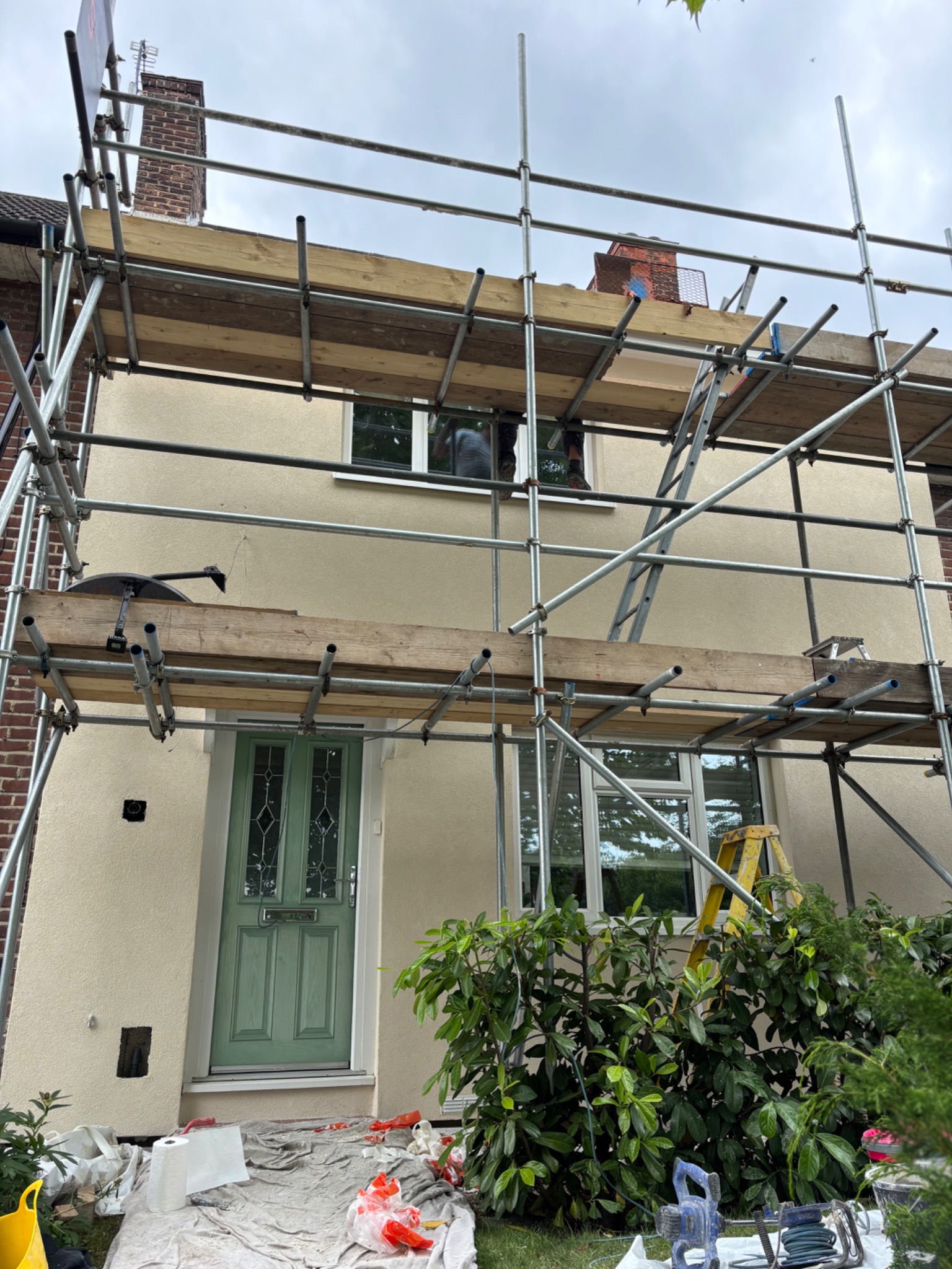 Dom jednorodzinny w rusztowaniach po nałożeniu beżowego tynku. Widoczna zielona stolarka okienna i drzwiowa. Prace elewacyjne w toku, osłonięte roślinnością.