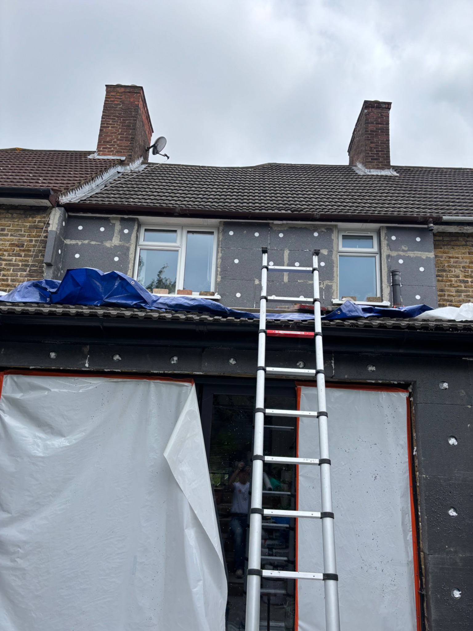 Ocieplenie elewacji domu jednorodzinnego z użyciem styropianu grafitowego. Widoczna drabina oparta o ścianę, zabezpieczona folią okienna stolarka. Etap przed nałożeniem tynku.