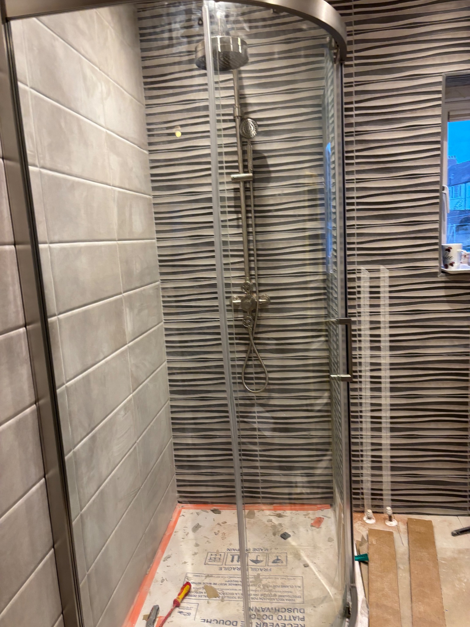 Nowoczesna kabina prysznicowa z chromowaną armaturą i szklanymi drzwiami, wykończona płytkami w odcieniach szarości z unikalnym wzorem fal. Widoczne narzędzia na podłodze.