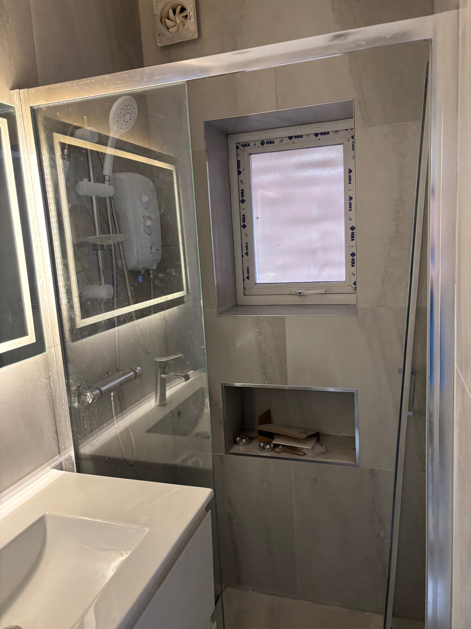 Wykończona łazienka w szarościach: kabina prysznicowa ze szklaną ścianą, umywalka z szafką, małe okno PCV i wnęka w ścianie na akcesoria. Widoczny wentylator.