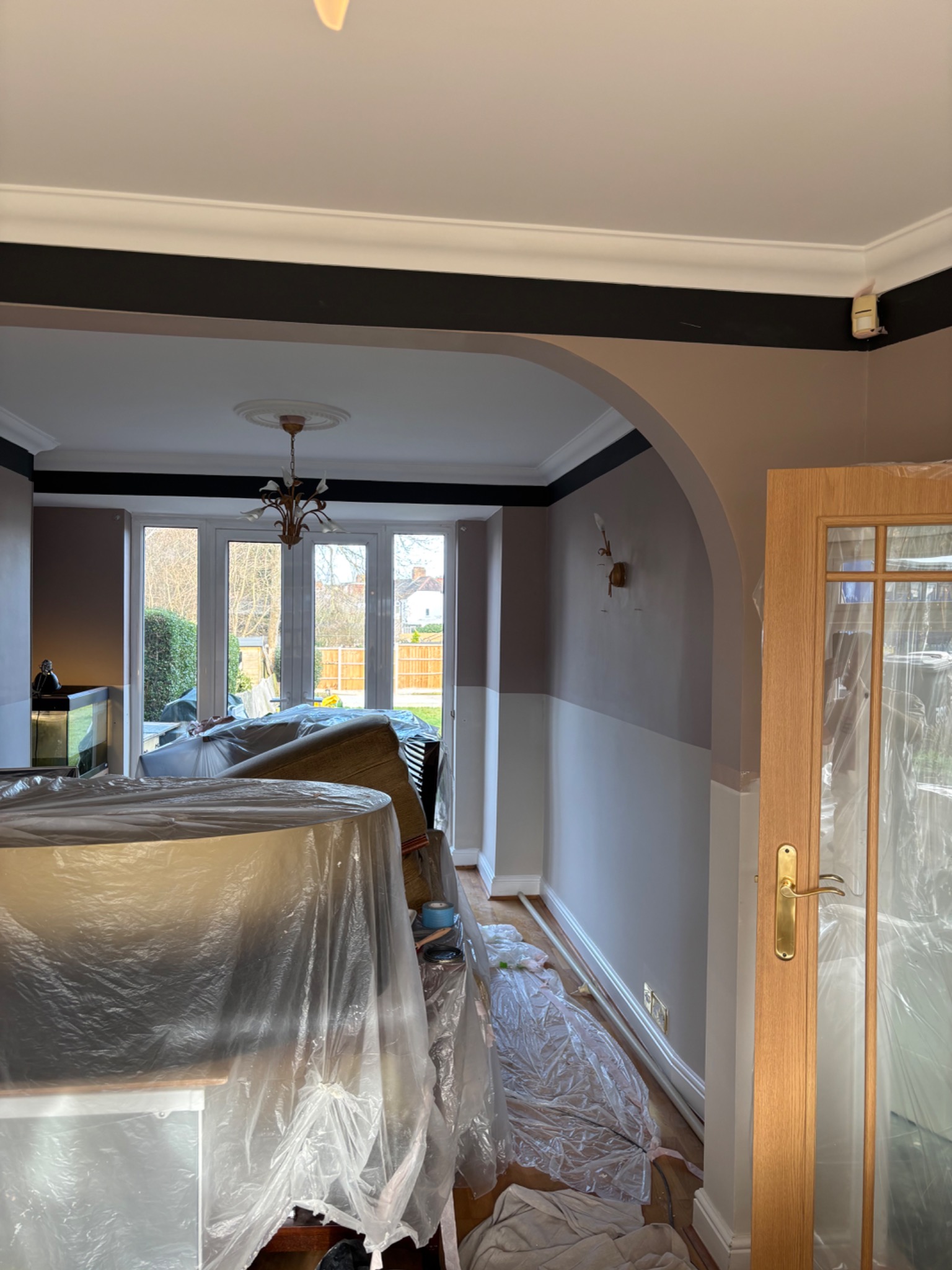 Pokój w trakcie malowania: meble zabezpieczone folią, ściany częściowo pomalowane na szaro i biało, widoczne listwy przypodłogowe i drzwi. Przygotowania do malowania.