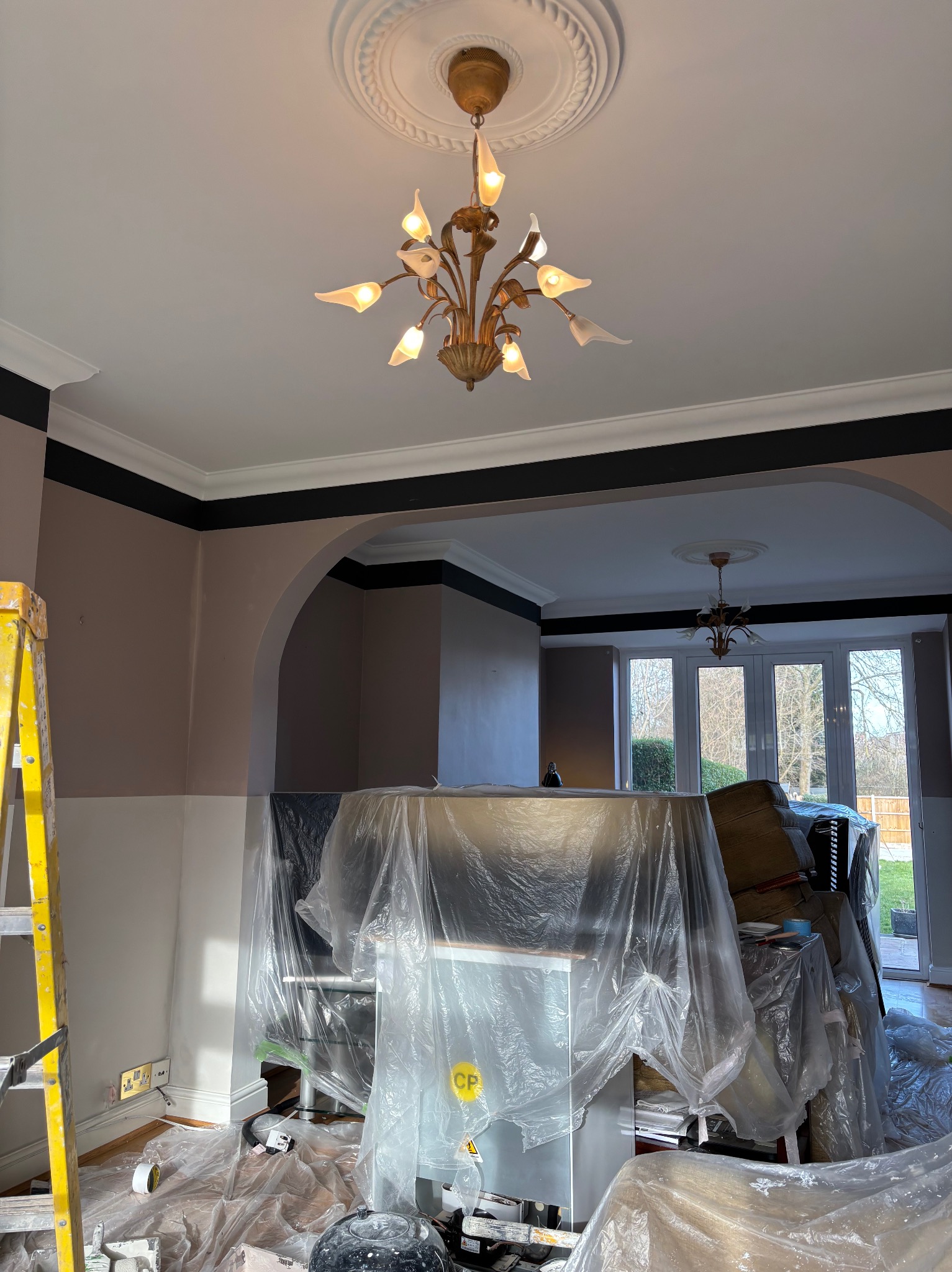 Wnętrze w trakcie malowania: ściany dwukolorowe (beż i biel), zabezpieczone meble folią, drabina, widoczny fragment sufitu z ozdobnym żyrandolem.