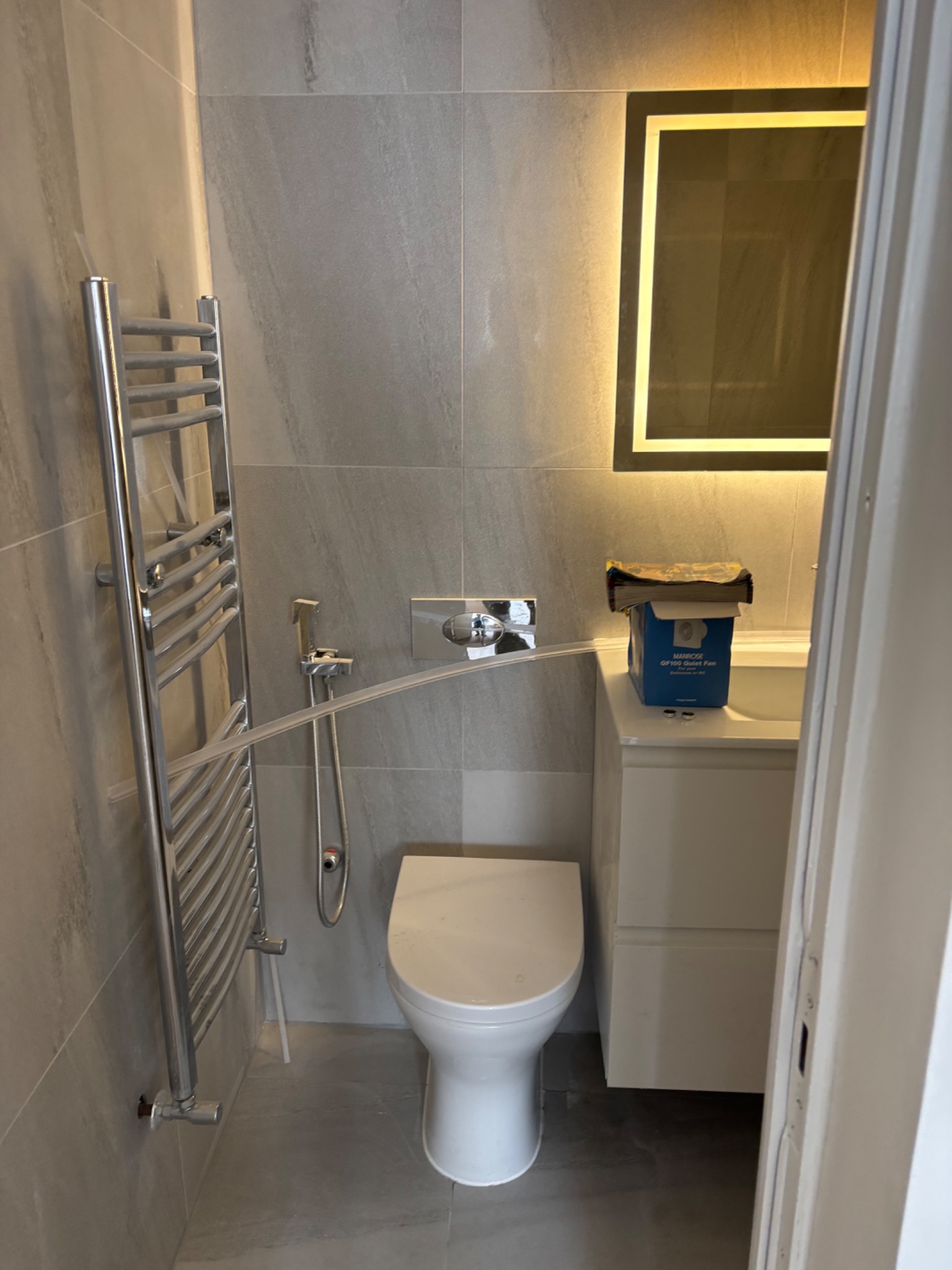 Wykończona łazienka z szarymi płytkami, podświetlanym lustrem, grzejnikiem drabinkowym i toaletą. Widoczny bidet i pudełko na szafce. Ujęcie z perspektywy drzwi.