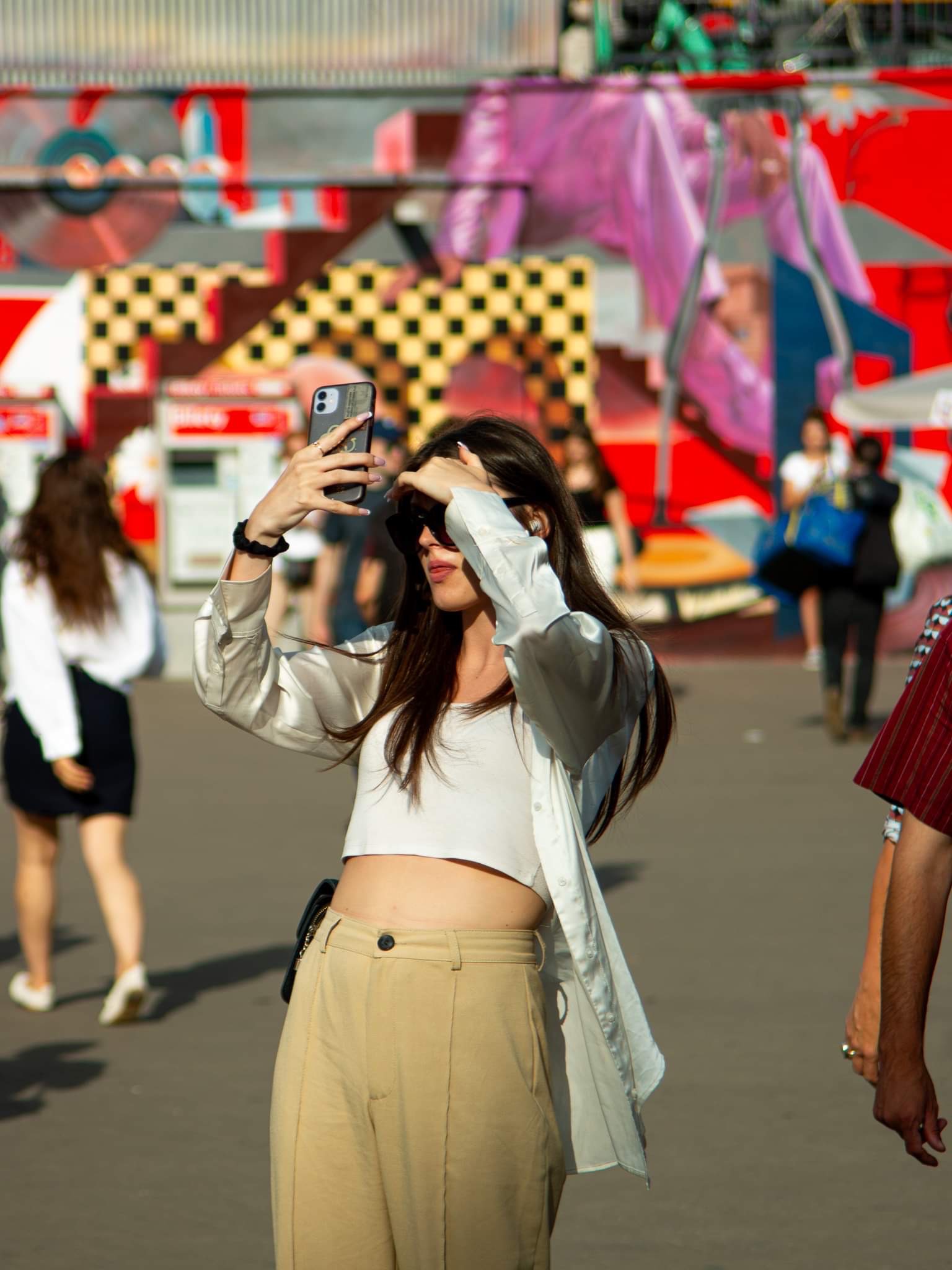 Kobieta w okularach przeciwsłonecznych robi zdjęcie telefonem na tle kolorowego muralu w przestrzeni miejskiej.