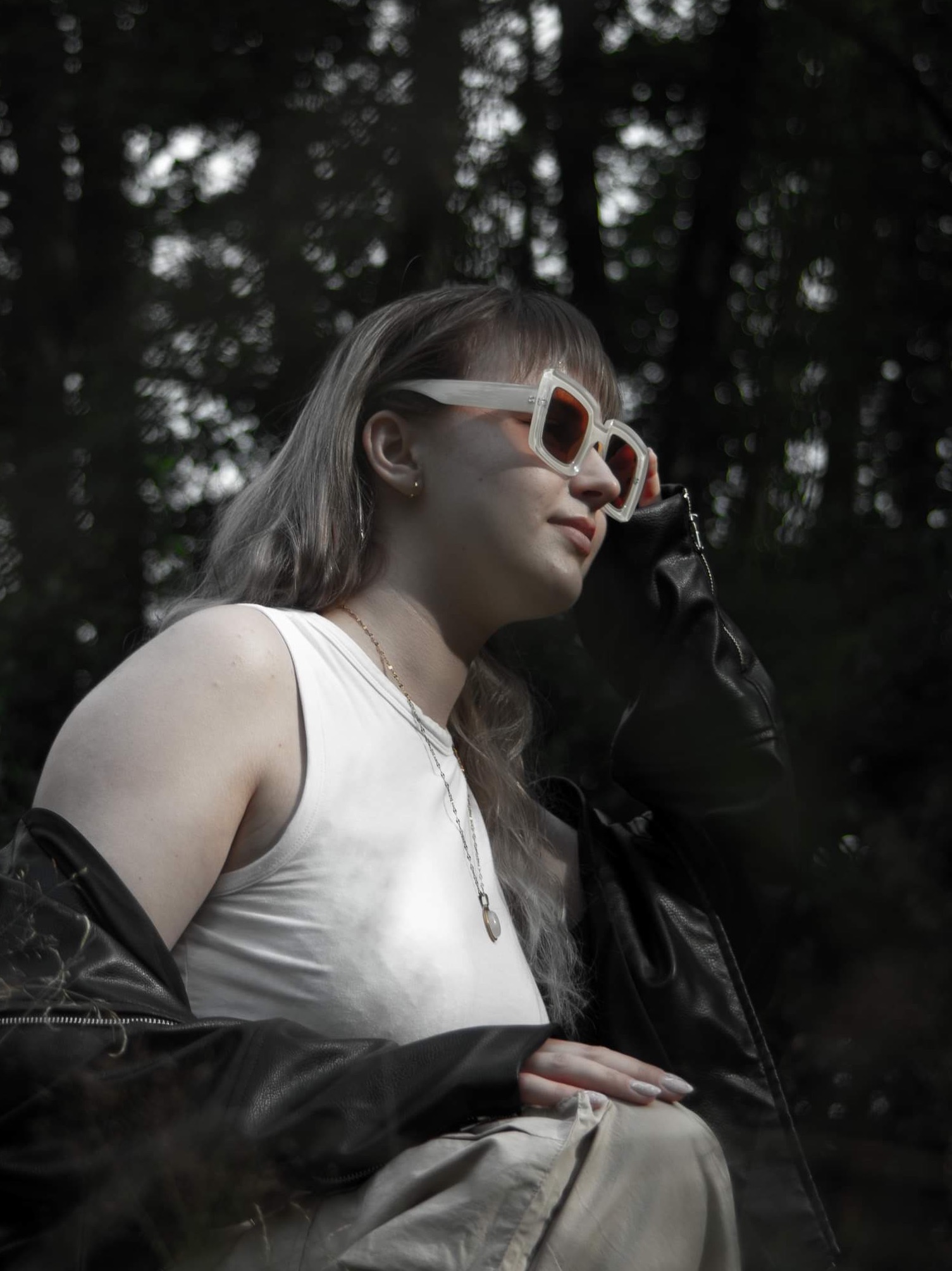 Portret kobiety w okularach przeciwsłonecznych, białym topie i skórzanej kurtce na tle rozmytego lasu, z delikatnym uśmiechem i dłonią przy okularach.