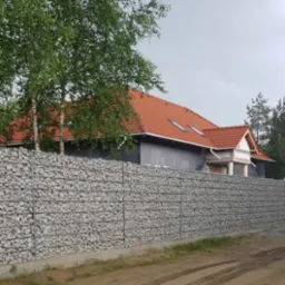 Długi, niski mur z gabionów wypełnionych jasnymi kamieniami, oddzielający budowę domu z czerwoną dachówką od polnej drogi z widocznymi śladami opon.