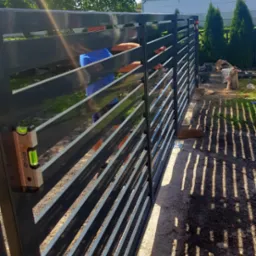 Montaż przęseł metalowego ogrodzenia panelowego z użyciem poziomicy, widoczne cienie na ziemi i elementy krajobrazu w tle.