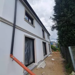 Boczny widok domu z białą elewacją, czarną rynną i beżowymi elementami dekoracyjnymi okien. Okna zabezpieczone folią, pomarańczowa rura odprowadzająca wodę leży na ziemi. Widoczne ślady prac...