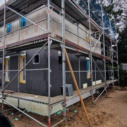 Usługi budowlane Grzegorz Junko - Dom w trakcie prac elewacyjnych, widoczne rusztowania, izolacja termiczna ścian zewnętrznych w kolorze czarnym oraz częściowo wykonana elewacja z białych bloczków.