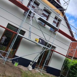 Usługi budowlane Grzegorz Junko - Malowanie elewacji domu jednorodzinnego z użyciem rusztowania, widoczny pracownik w kombinezonie ochronnym, częściowo otynkowana ściana z oknami z brązowymi ramami.