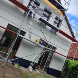 Malowanie elewacji domu jednorodzinnego z użyciem rusztowania, widoczny pracownik w kombinezonie ochronnym, częściowo otynkowana ściana z oknami z brązowymi ramami.