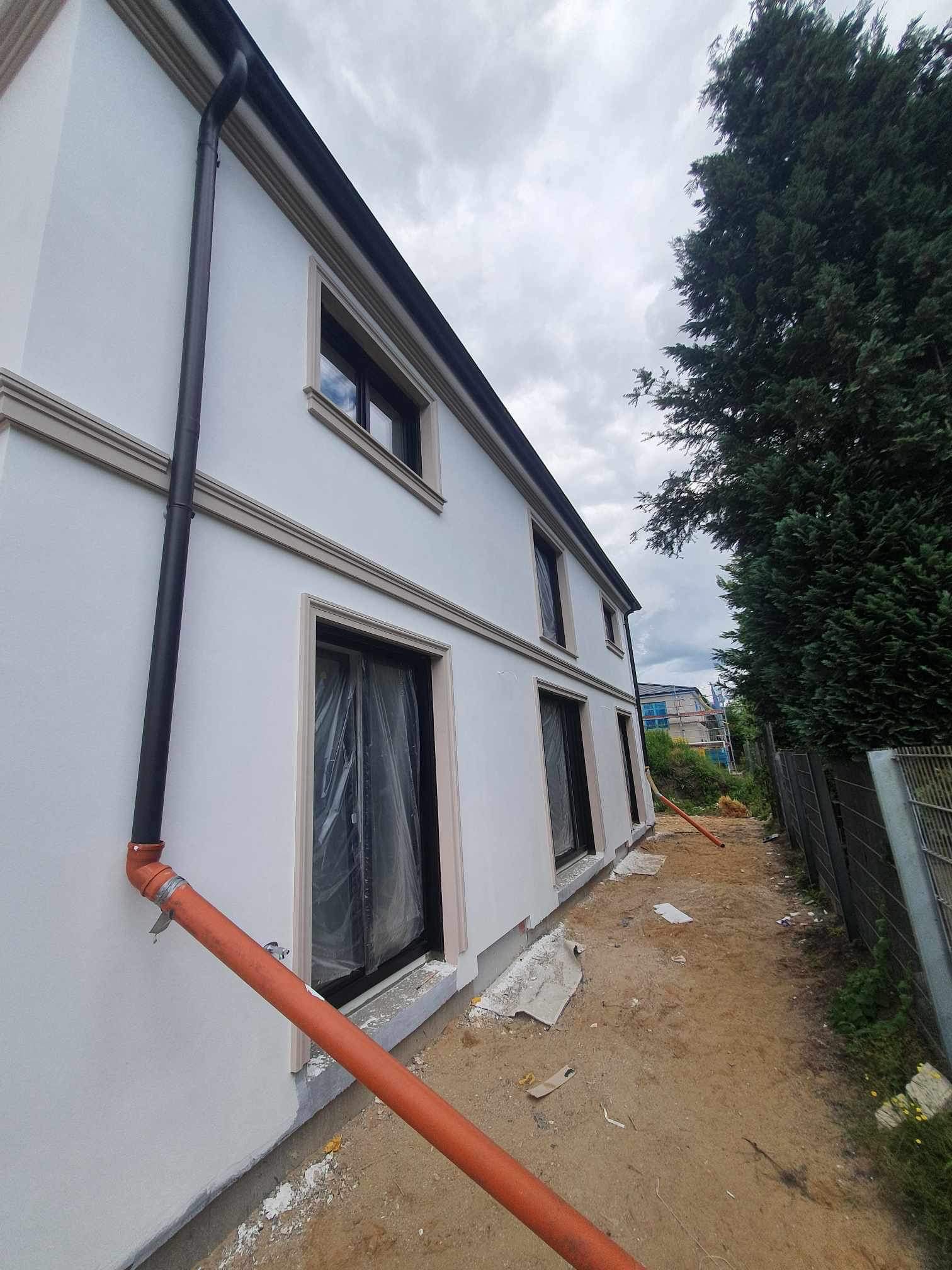 Boczny widok domu z białą elewacją, czarną rynną i beżowymi elementami dekoracyjnymi okien. Okna zabezpieczone folią, pomarańczowa rura odprowadzająca wodę leży na ziemi. Widoczne ślady prac...