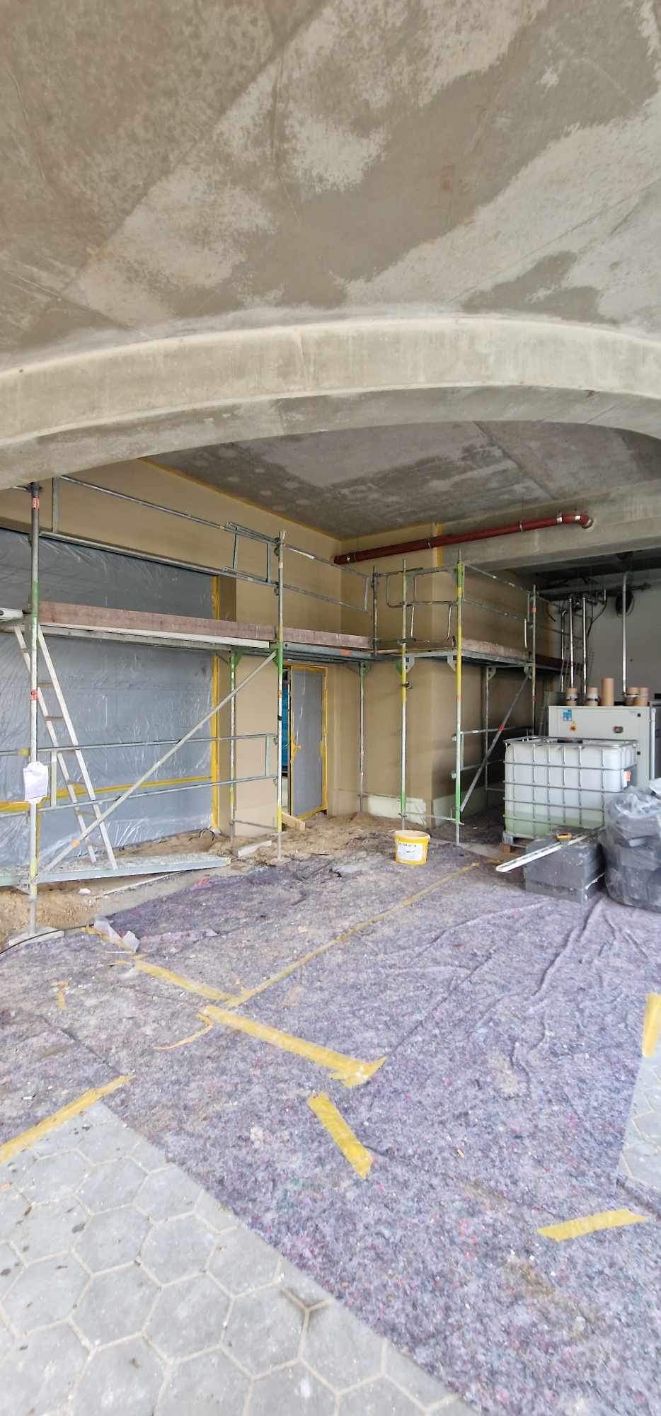 Wnętrze w trakcie prac budowlanych z rusztowaniem, zabezpieczoną podłogą, ścianami przygotowanymi do wykończenia i widocznymi instalacjami.