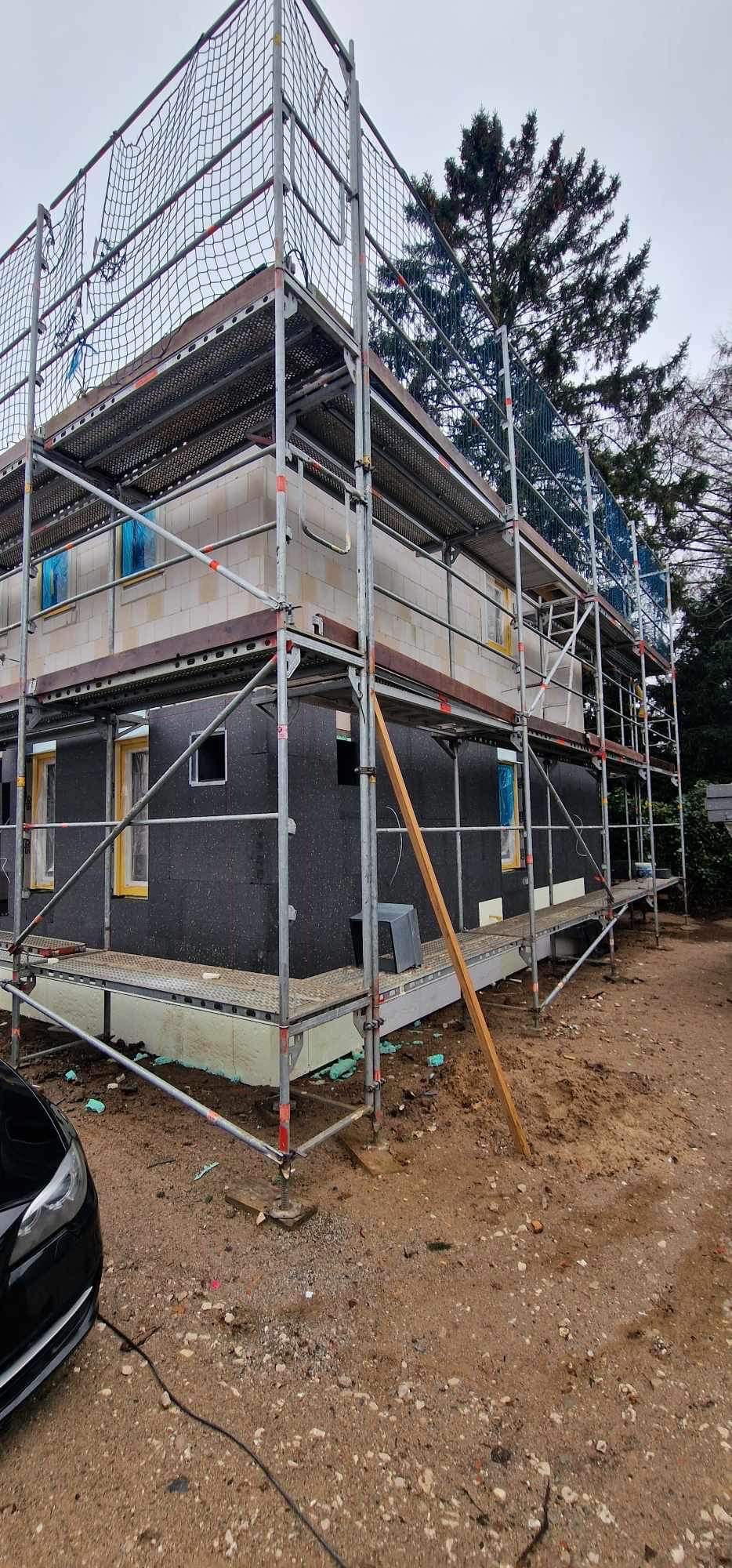 Dom w trakcie prac elewacyjnych, widoczne rusztowania, izolacja termiczna ścian zewnętrznych w kolorze czarnym oraz częściowo wykonana elewacja z białych bloczków.