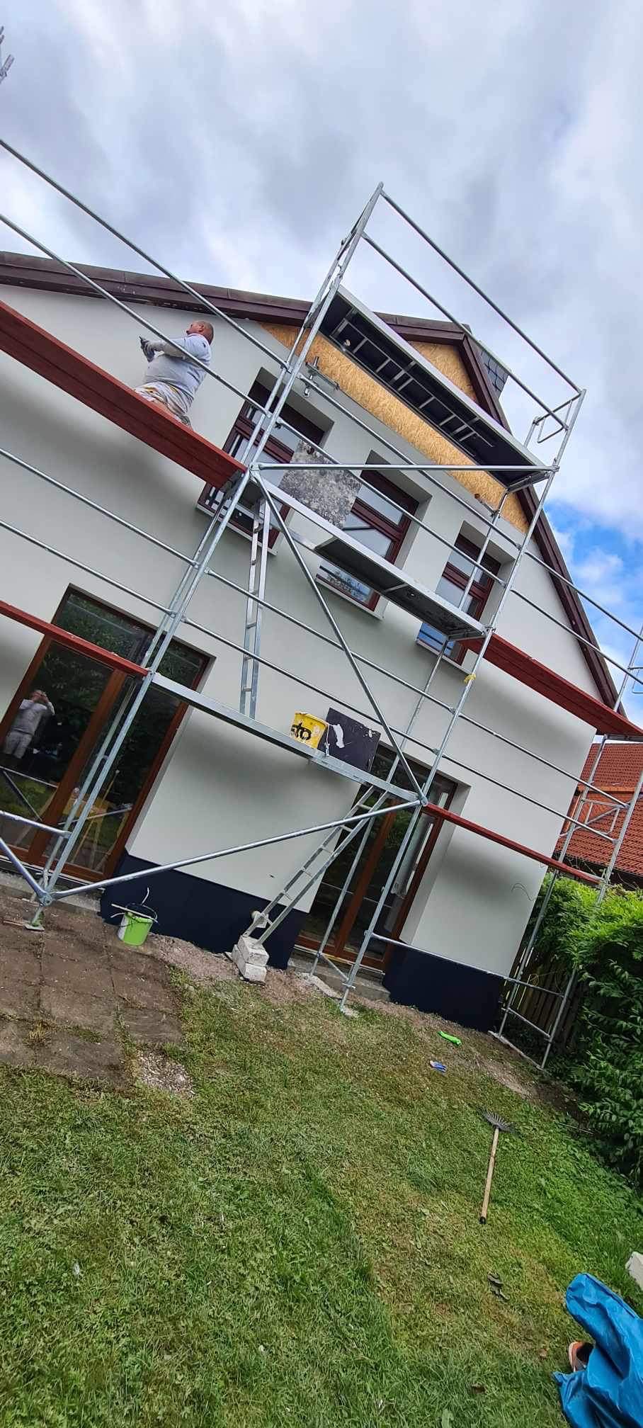 Malowanie elewacji domu jednorodzinnego z użyciem rusztowania, widoczny pracownik w kombinezonie ochronnym, częściowo otynkowana ściana z oknami z brązowymi ramami.
