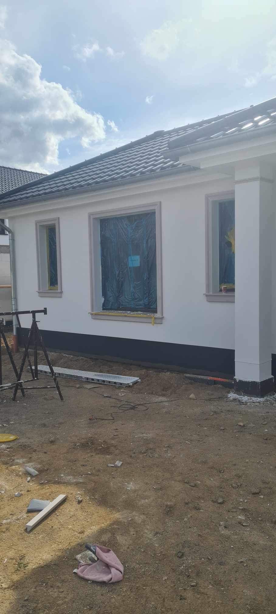 Fragment elewacji domu z oknami zabezpieczonymi folią malarską, świeżo otynkowany cokół, widoczne rusztowanie i teren budowy.