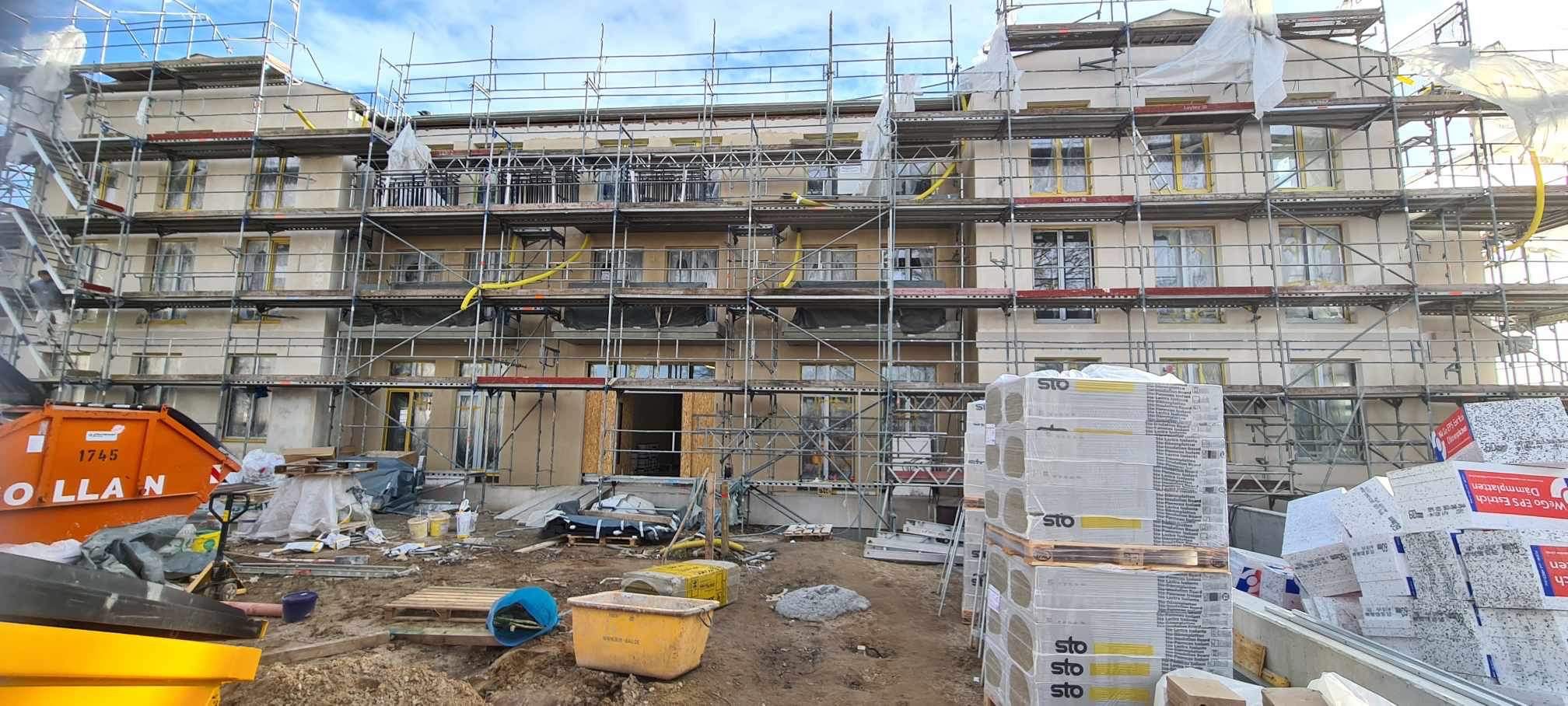 Wielopiętrowy budynek w trakcie prac elewacyjnych, otoczony rusztowaniami, z widocznymi materiałami izolacyjnymi STO na paletach i pomarańczowym kontenerem na gruz w tle.