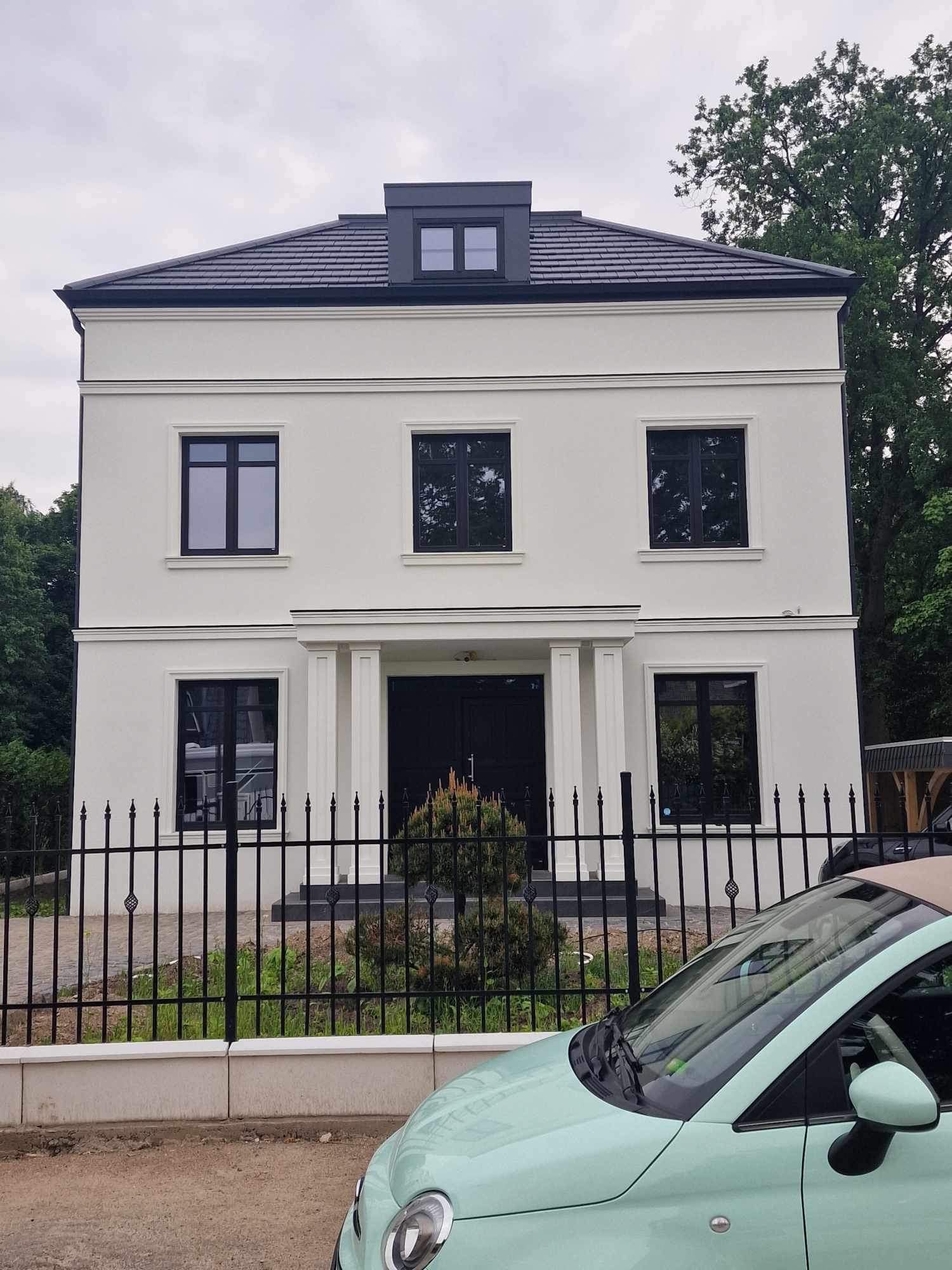Dwupiętrowy dom z białą elewacją, czarną stolarką okienną i ciemnoszarą dachówką, ogrodzony czarnym metalowym płotem, z zielonym samochodem na pierwszym planie.