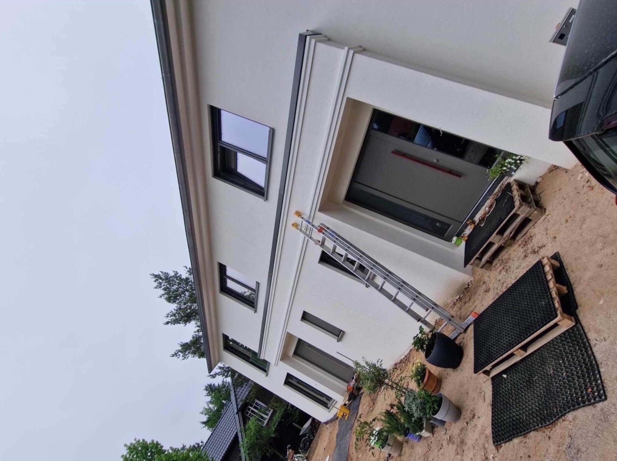 Nowoczesny dom jednorodzinny w Kętrzynie po świeżym tynkowaniu elewacji, z aluminiową drabiną opartą o ścianę i paletami zabezpieczającymi teren wokół wejścia.