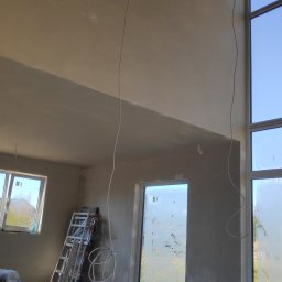 Home Future Chojnowski Andrzej - Wysokie wnętrze w trakcie prac wykończeniowych, ściany pokryte świeżym tynkiem, widoczne okna, drabina i wystające kable elektryczne.