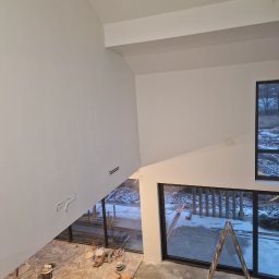 Home Future Chojnowski Andrzej - Wnętrze w trakcie prac wykończeniowych: świeżo pomalowane białe ściany w wysokim pomieszczeniu z dużymi oknami, drabina oparta o okno, widoczne puszki z farbą i narzędzia na podłodze zabezpieczonej...