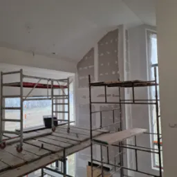 Wnętrze w trakcie prac wykończeniowych: widoczne rusztowania, ściany z fragmentami nałożonej zaprawy, okna zabezpieczone folią.