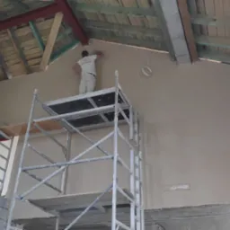 Pracownik na rusztowaniu aplikuje gładź na ścianę pod skośnym dachem, widoczne drewniane elementy konstrukcyjne, przewody elektryczne i fragmenty czerwonej ściany.